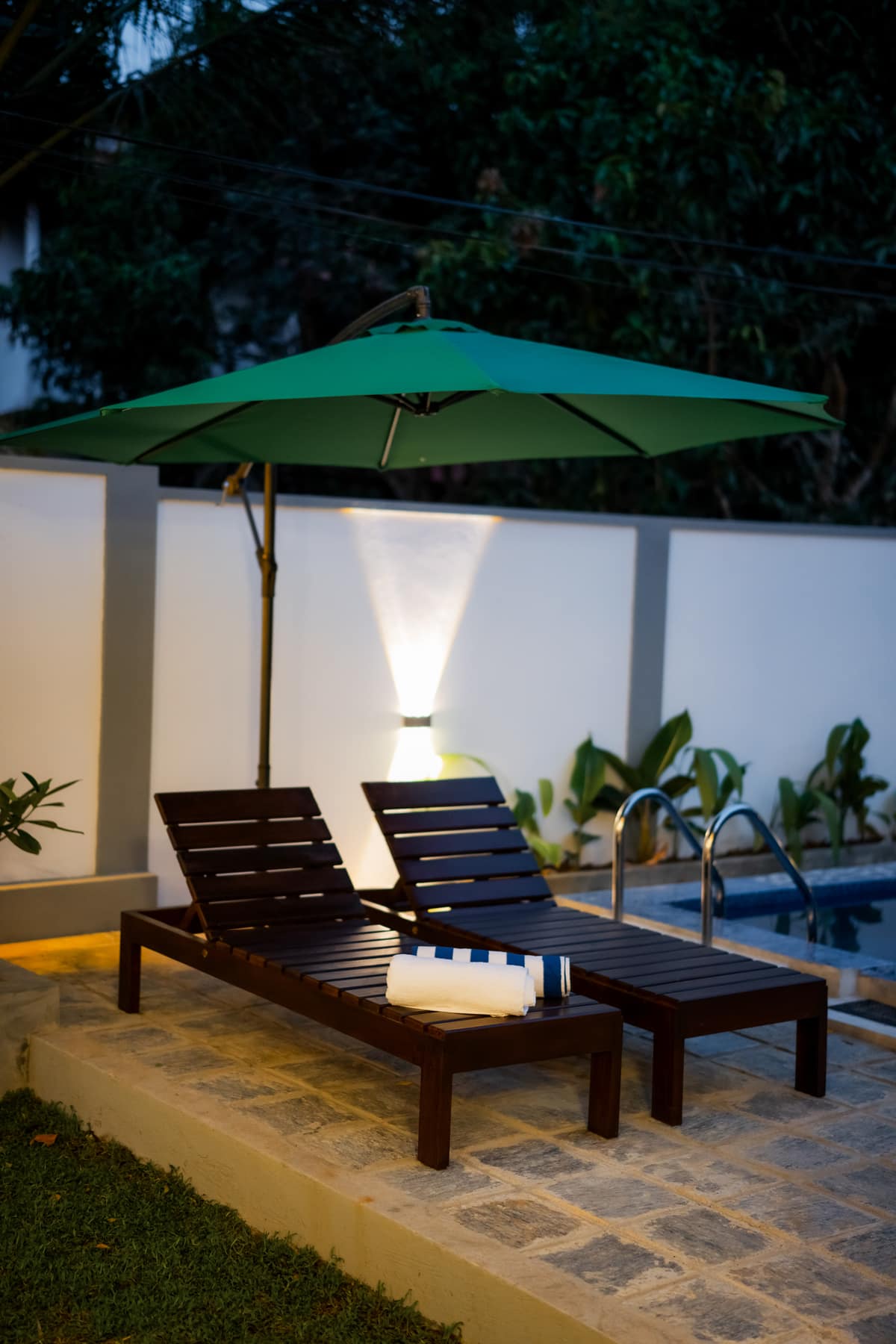 Two wooden loungers and a folded towel beside the lit pool at dusk
