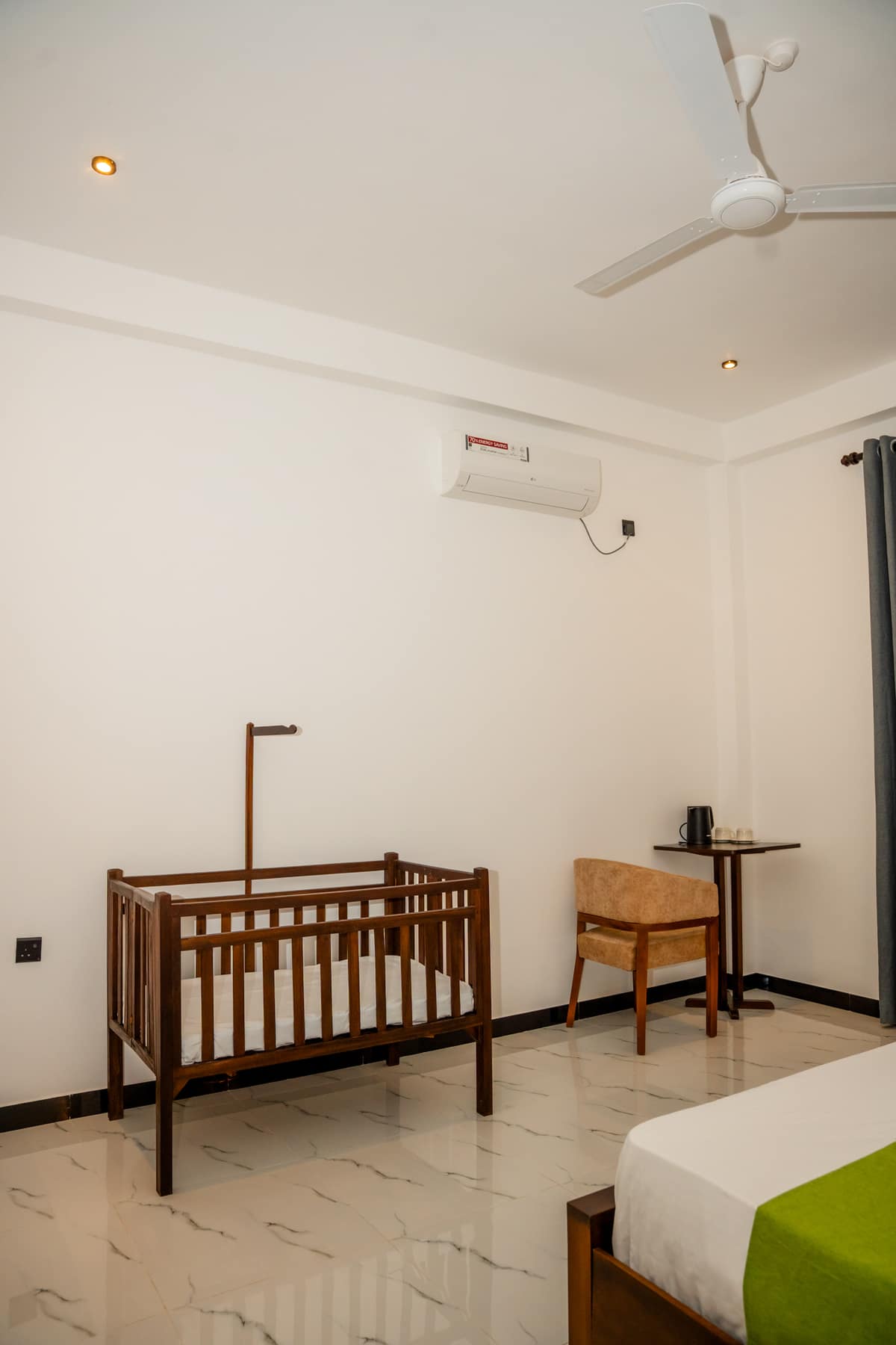 A wooden cot set up beside the bed, with a chair and a writing table against the wall
