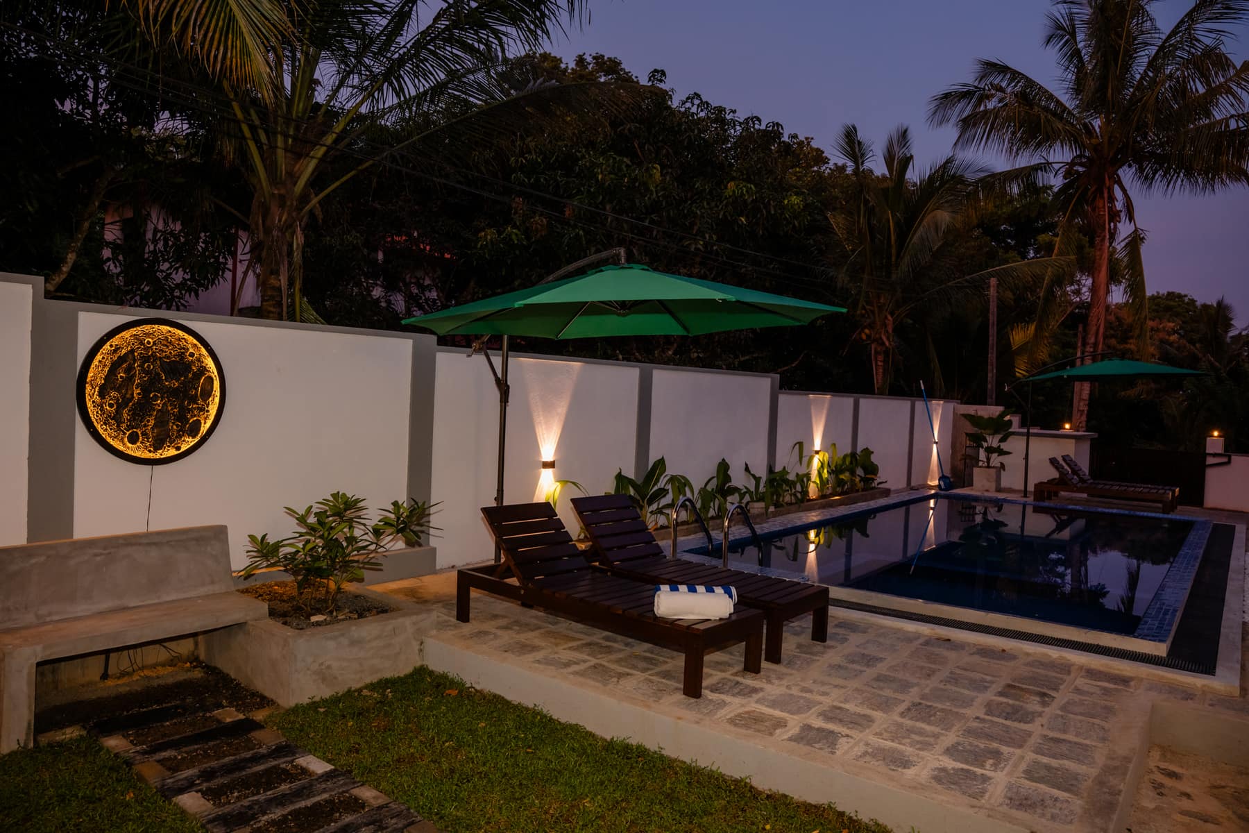 The poolside terrace at dusk, lit moon artwork on the garden wall and coconut palms behind it